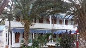 A two-story building with a blue awning over the entrance, surrounded by various plants and a wire fence. - Baia Verde (Tarrafal)