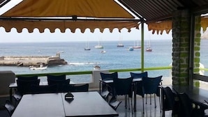 An outdoor dining area with tables and chairs, overlooking a coastal view with boats and a rocky shoreline. - Baia Verde (Tarrafal)