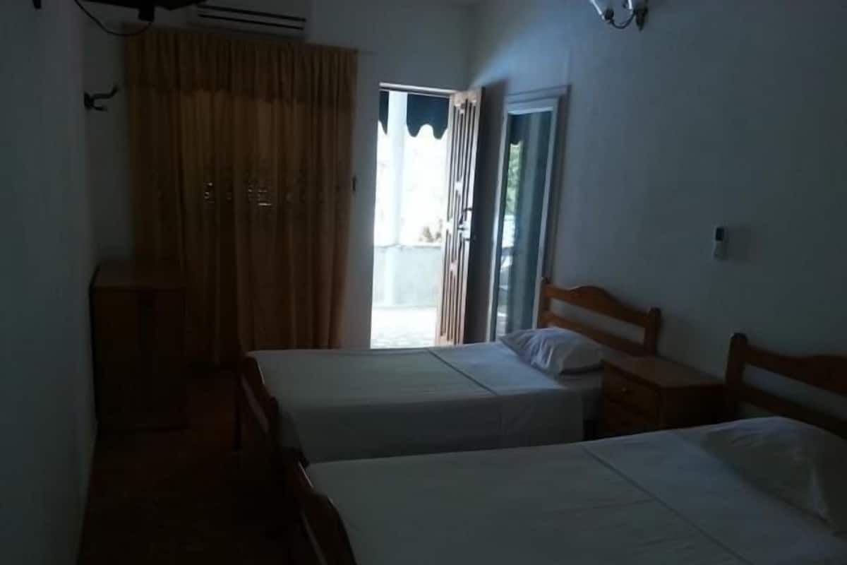 Ein Zimmer mit zwei Einzelbetten, einem Holznachttisch, einem kleinen Tisch und einem Fenster mit Vorhängen.