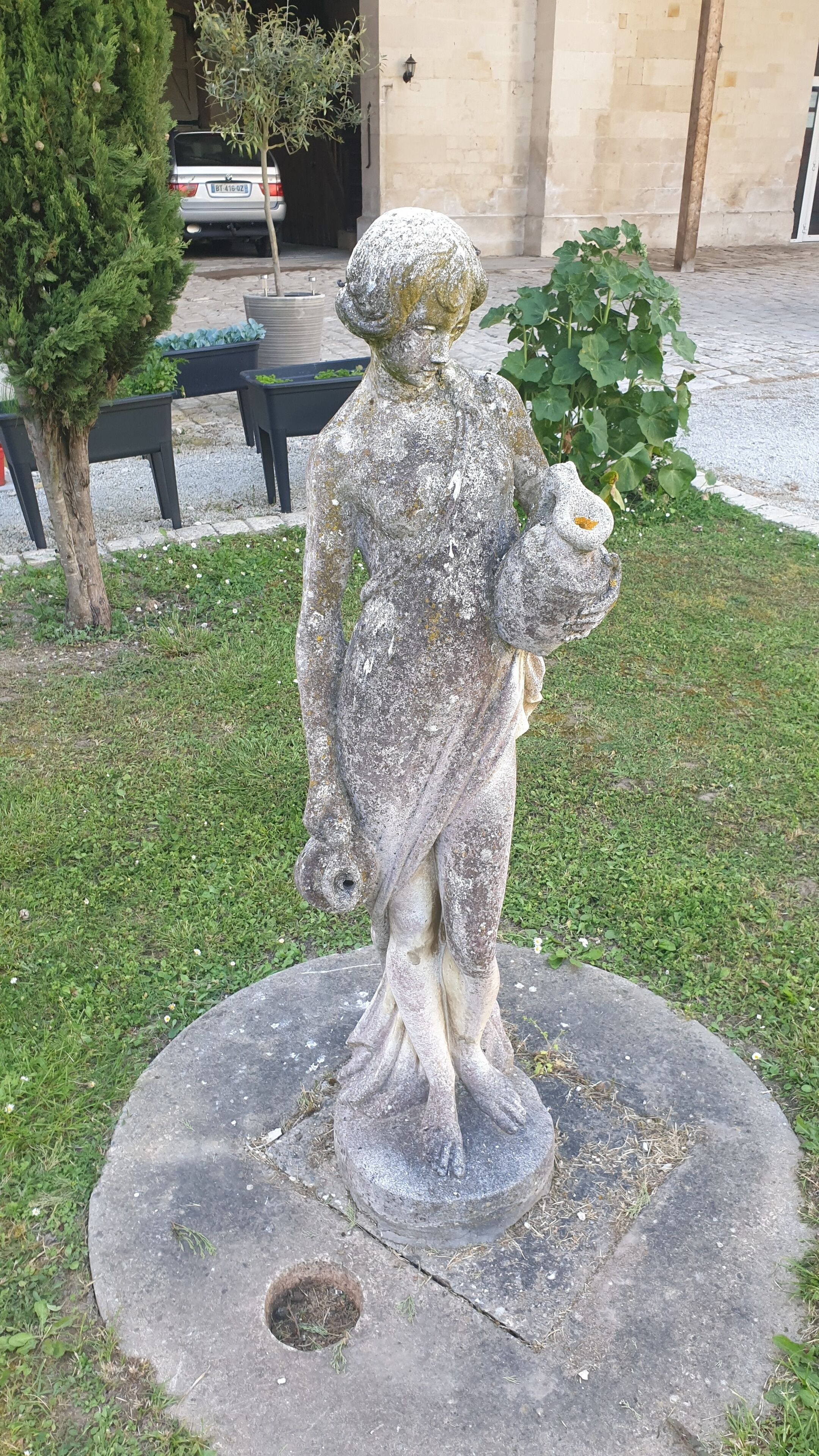 A weathered statue of a person holding a flower, set on a stone base in a grassy area with a building and parked cars in the background.