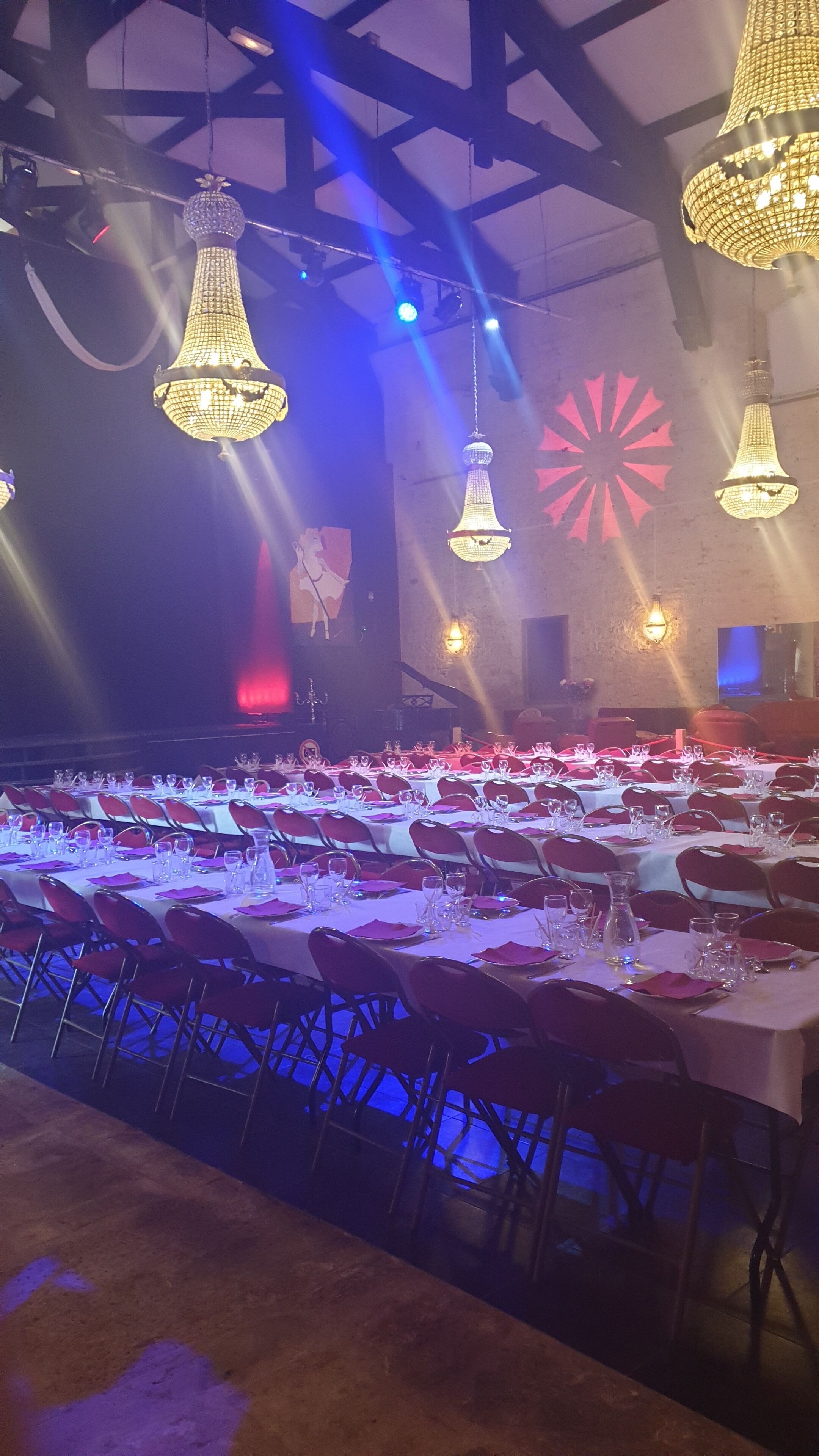 A banquet hall with multiple tables set for a formal event, featuring chandeliers and stage lighting.