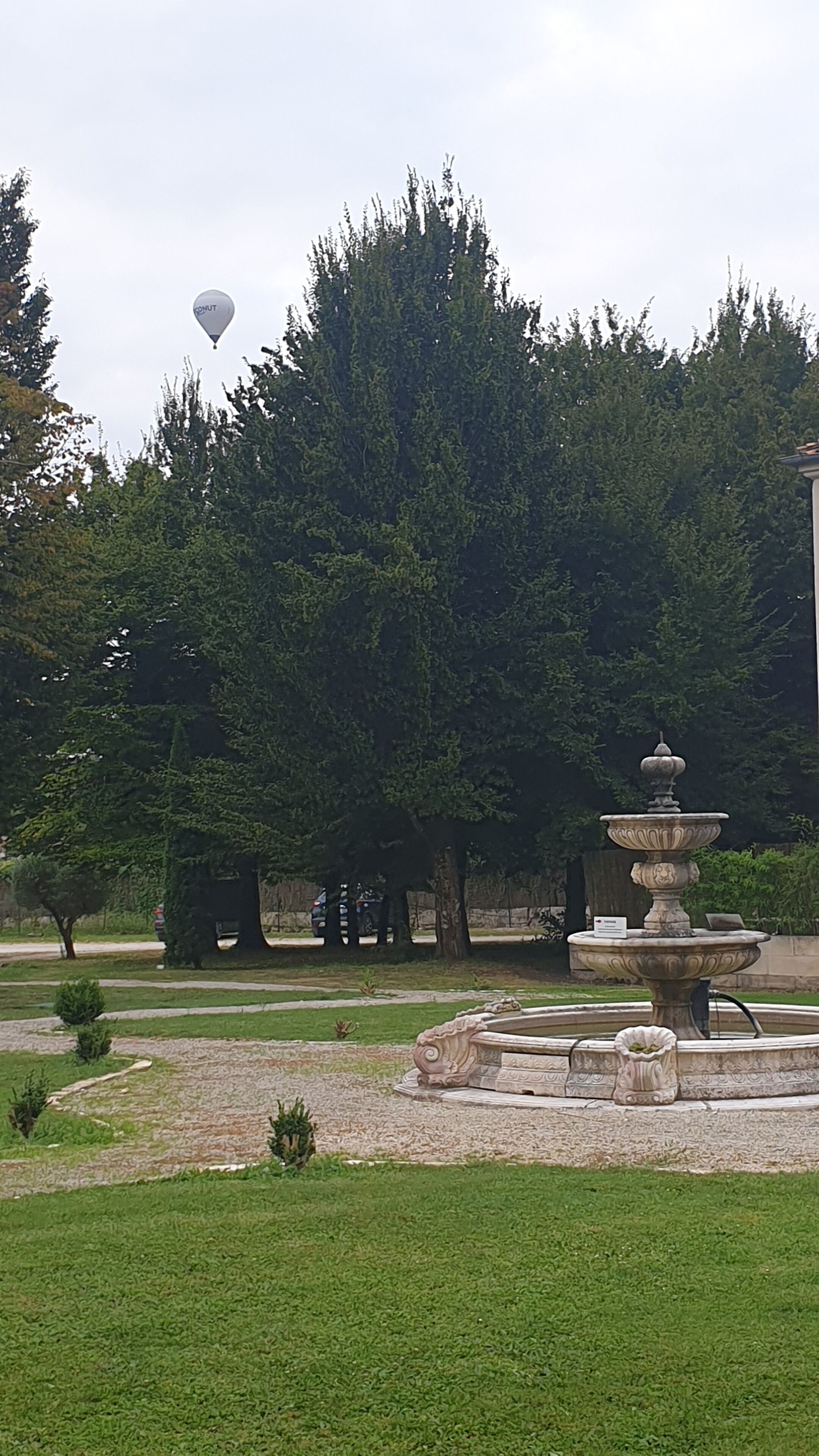A park with a fountain, trees, and a hot air balloon in the sky.