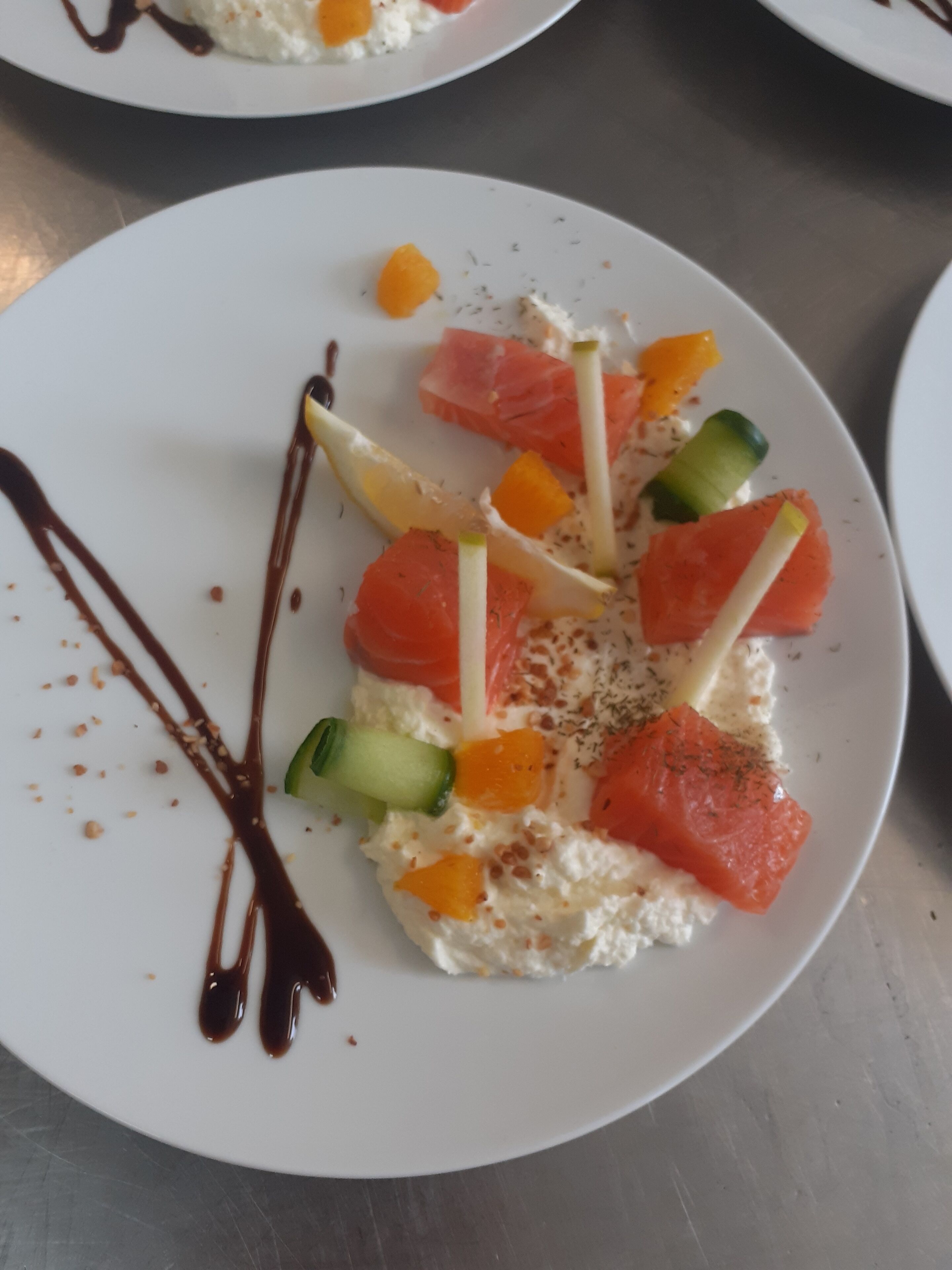 Un piatto bianco con un filetto di salmone, spicchi d'arancia, cetrioli e una base cremosa, guarnito con erbe aromatiche e un filo di cioccolato.