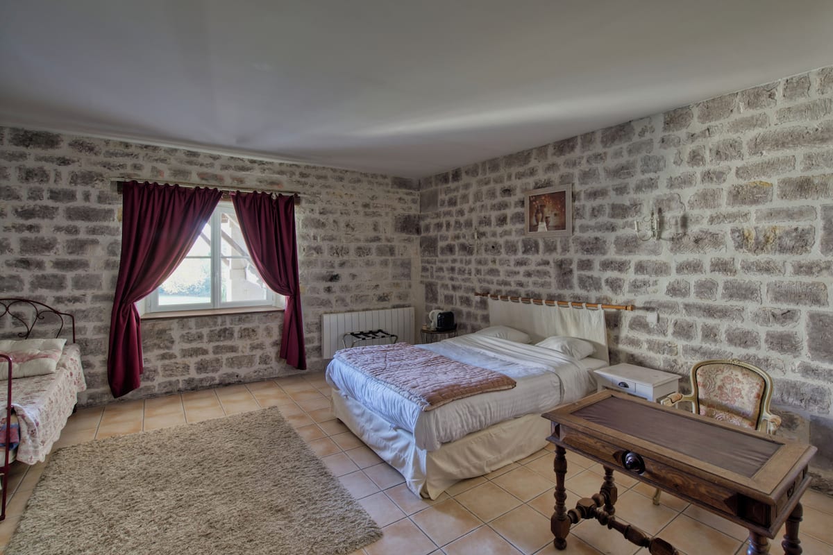 A bedroom with a stone wall, a bed with a white headboard, a wooden nightstand, a chair, and a window with curtains.
