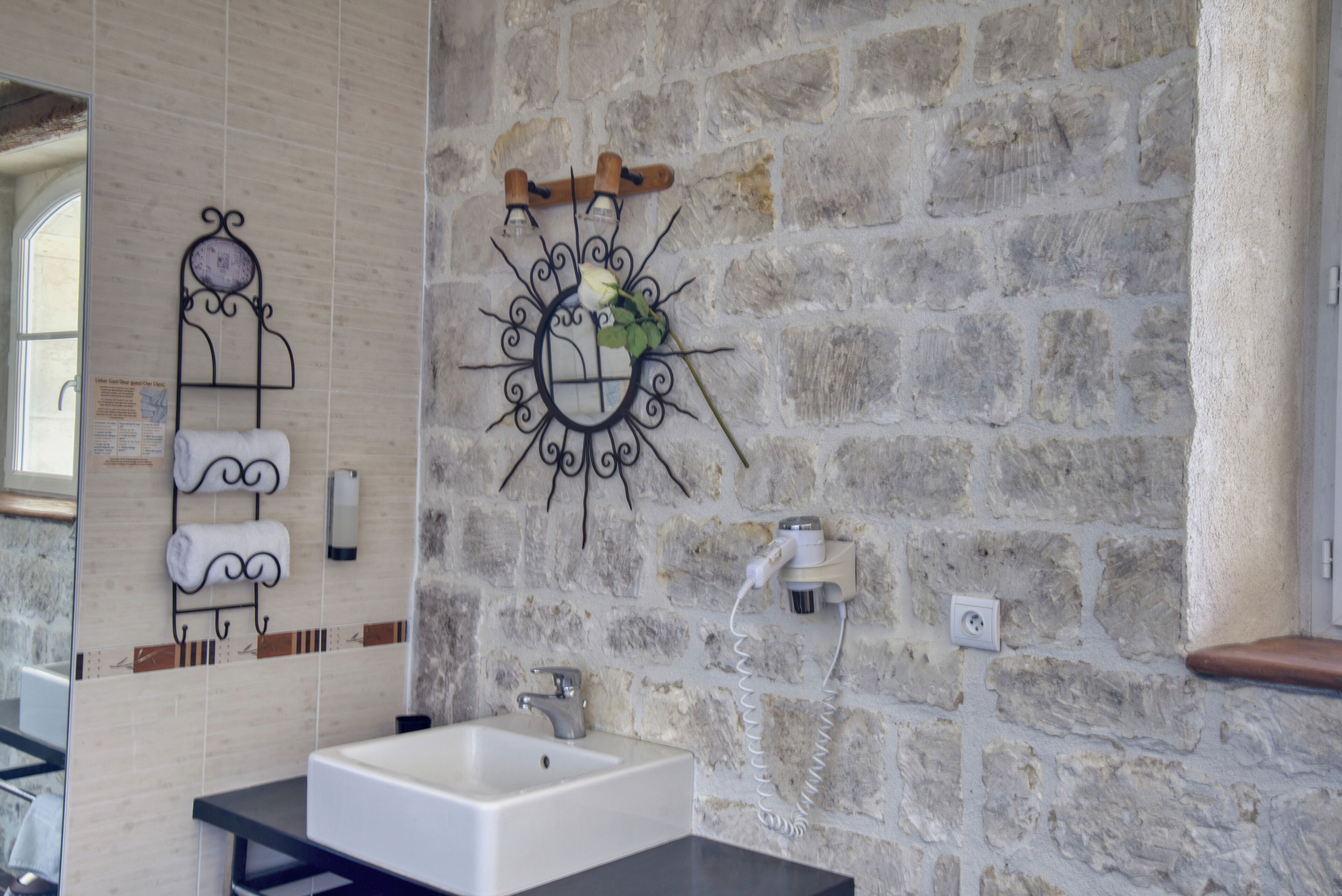 Une salle de bain avec un mur en pierre, un lavabo blanc et un miroir décoratif orné d’une rose.
