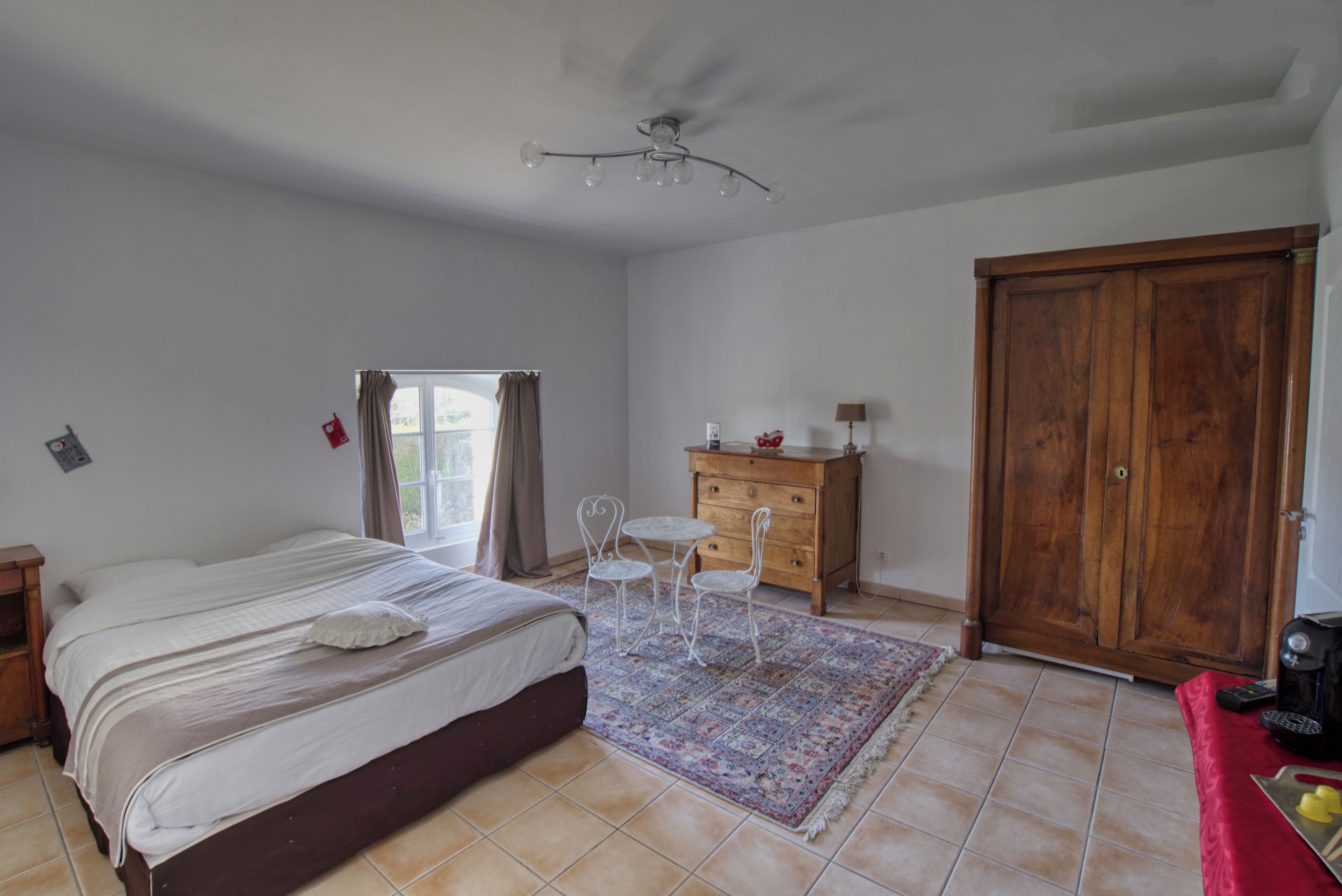 Une chambre à coucher comprenant un lit, une commode, une armoire, une petite table avec des chaises et une fenêtre avec des rideaux.