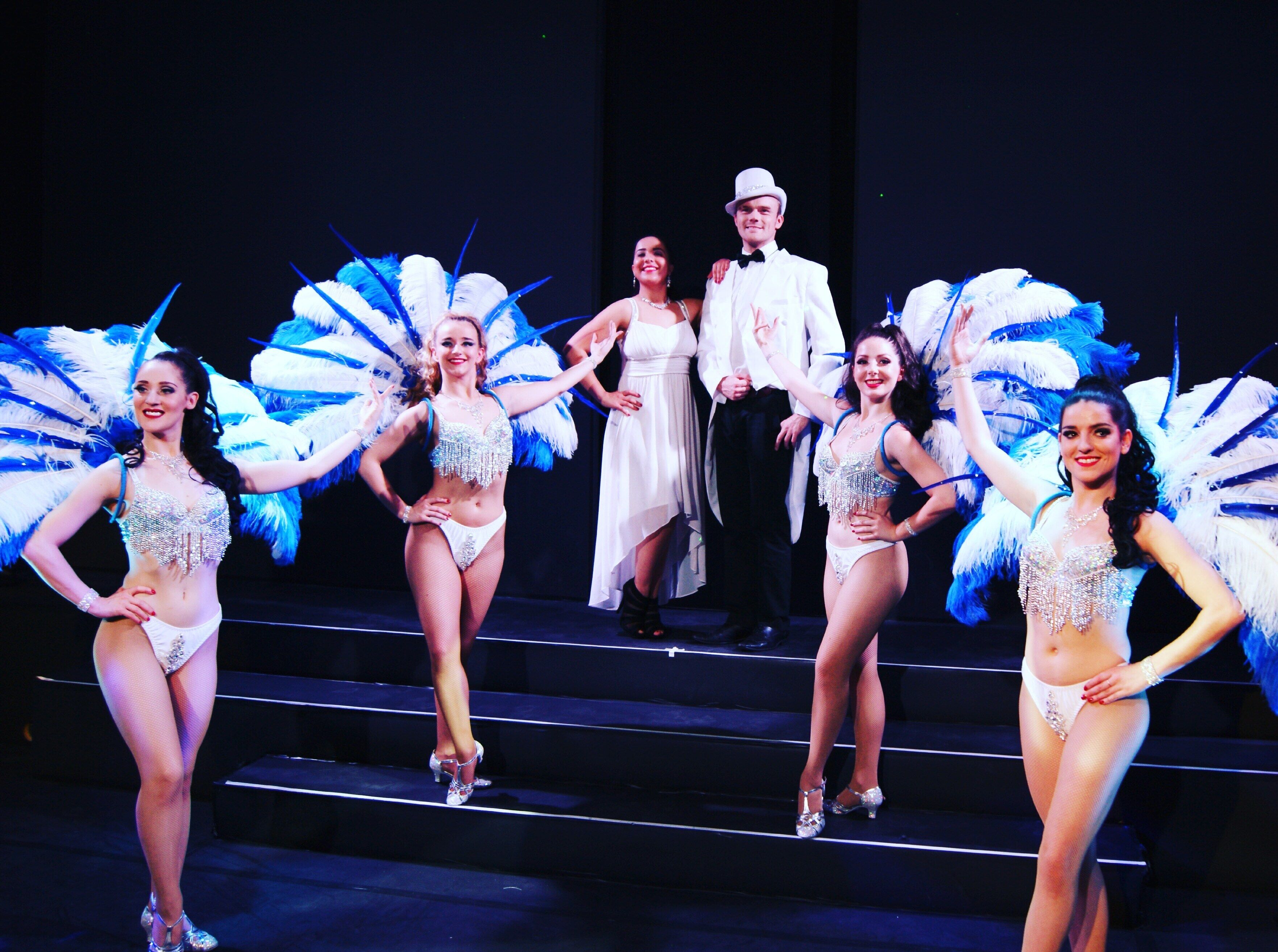 A group of performers on stage with feathered costumes and a man in a suit.