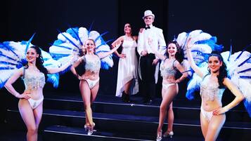 A group of performers on stage with feathered costumes and a man in a suit.
