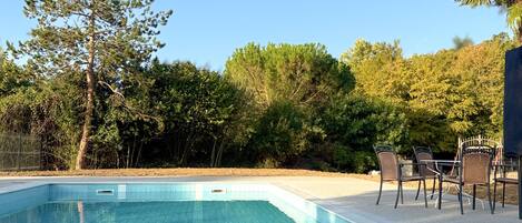 A swimming pool with tiled edges, surrounded by trees and a patio with chairs.