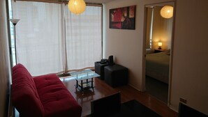 A modern living room with a red sofa, a glass coffee table, and a floor lamp. There is a painting on the wall and a large window with sheer curtains. - Departamentos Centro Urbano Providencia (Santiago)