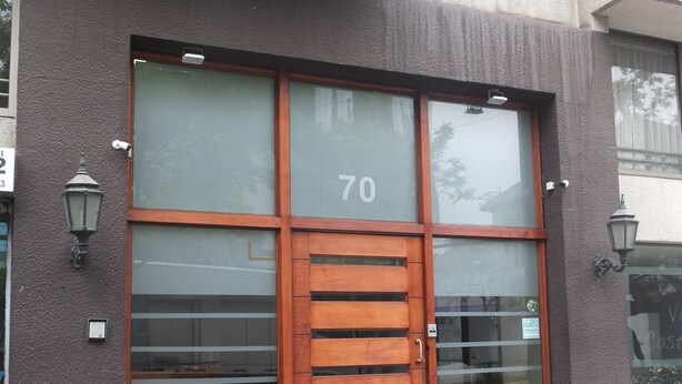 A building with a large glass facade, a wooden door, and two wall-mounted lamps. - Departamentos Centro Urbano Providencia (Santiago)