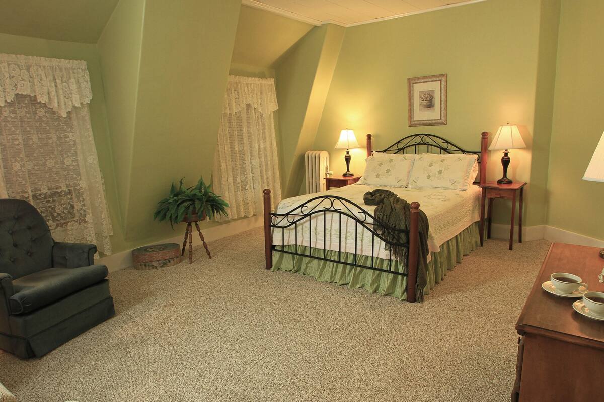 A bedroom with a bed, two bedside lamps, a chair, a small table with a lamp, and a potted plant.