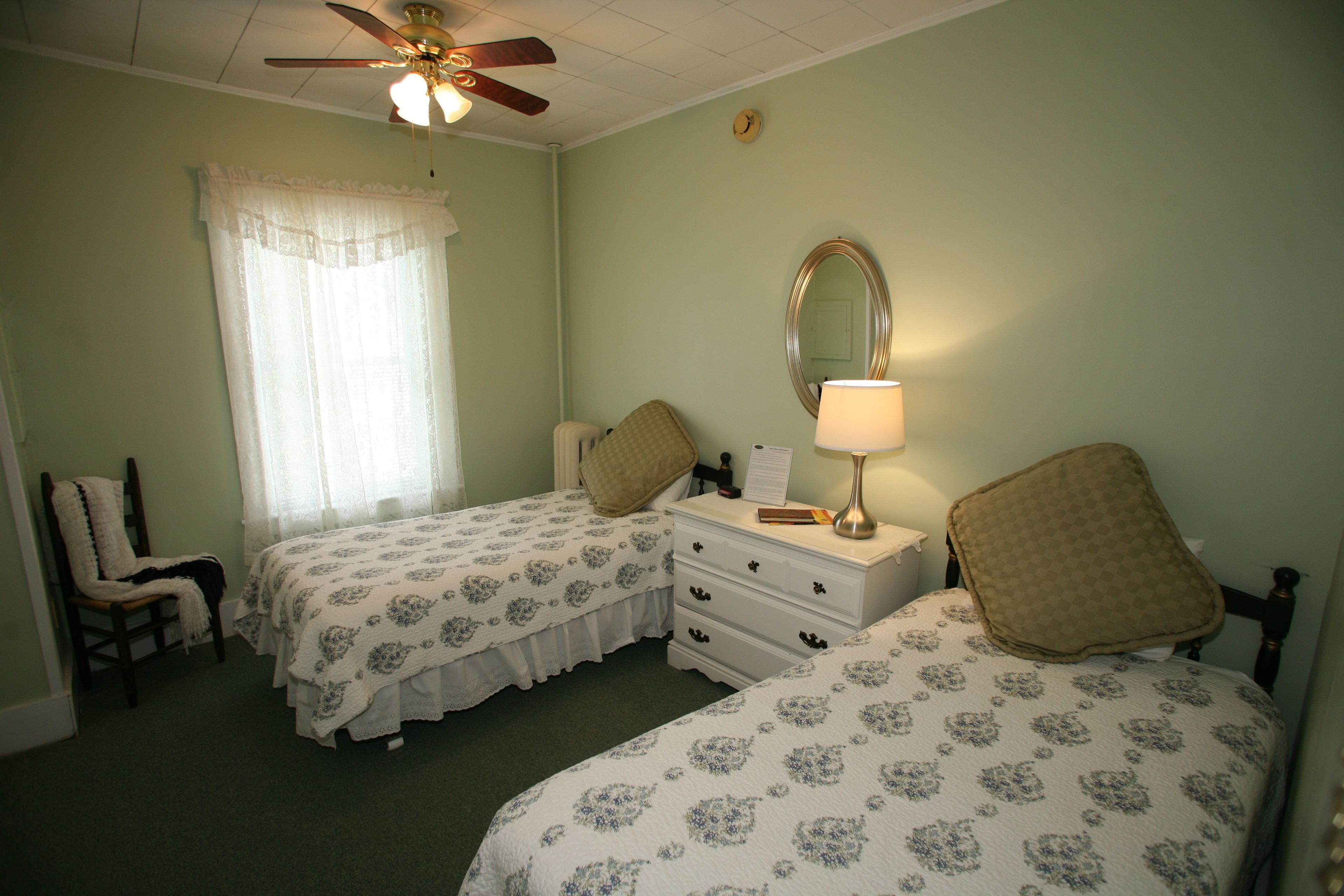 A bedroom with two beds, a ceiling fan, a window with curtains, a bedside table with a lamp, and a chair.