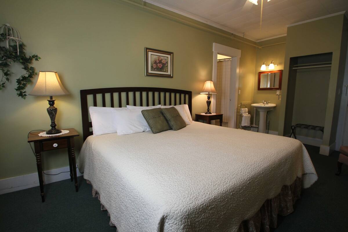 A neatly made bed with a white quilt, two bedside tables with lamps, a framed picture on the wall, and a bathroom with a sink and mirror.