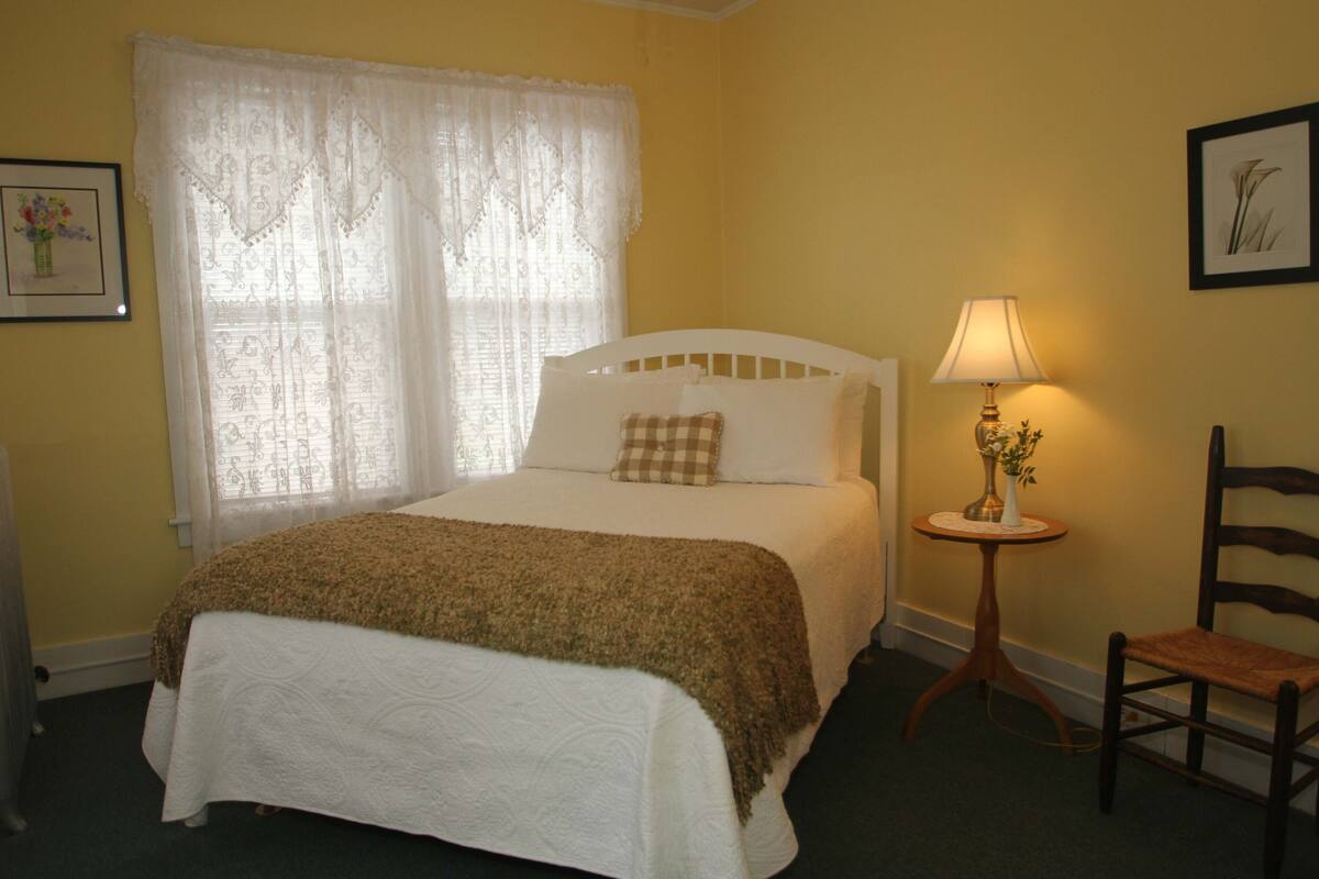 A neatly made bed with a white headboard, a small bedside table with a lamp, a chair, and a window with sheer curtains.