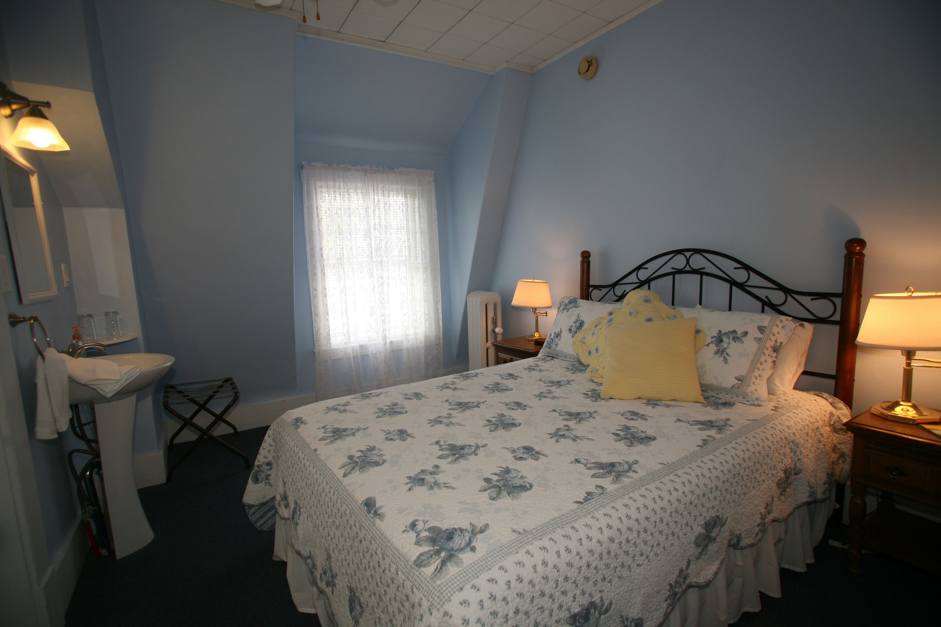 Una camera da letto con un letto, due comodini, un tavolo da notte, una finestra con tende, una sveglia a muro e un bagno con lavandino e specchio.