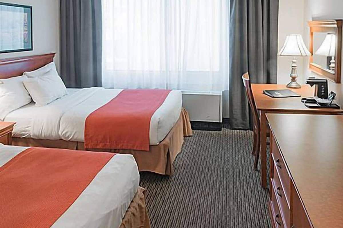 Hotellihuone, jossa on kaksi sänkyä, ty öpöytä, tuoli, lamppu ja suuri ikkuna, jossa on verhot.