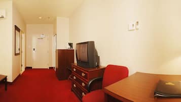 A hotel room with a red carpet, a sofa, a desk with a lamp, a television, a bed with a patterned bedspread, and a window with curtains.