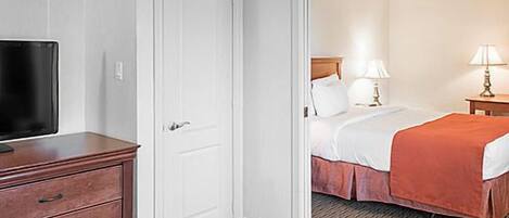 A hotel room with a wooden dresser, a television, a bed with a red blanket, and a nightstand with a lamp.
