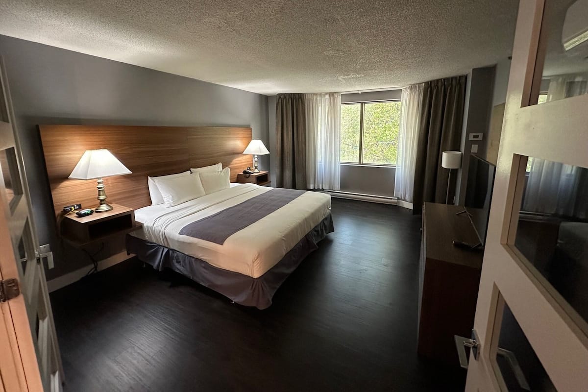 Habitación de hotel con una cama grande, dos mesitas de noche con lámparas, una ventana con cortinas y una puerta.