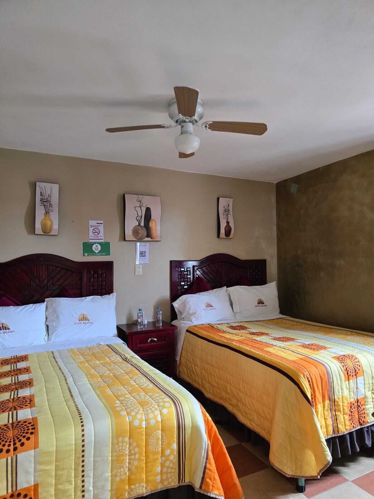 A hotel room with two beds, a ceiling fan, artwork on the walls, and a nightstand with a water bottle.