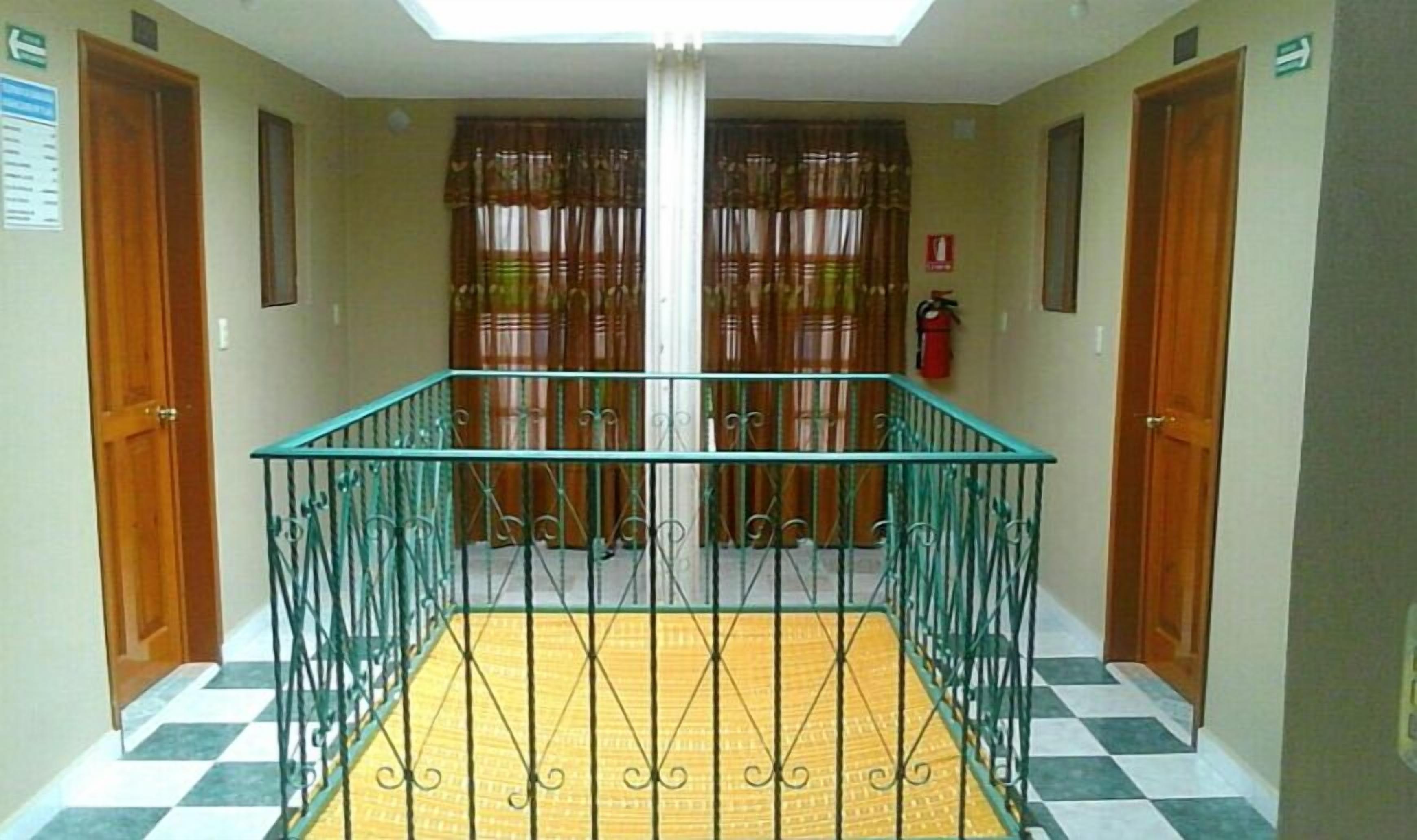 Pasillo de hotel con barandilla de vidrio central, suelo a cuadros y puertas de madera.