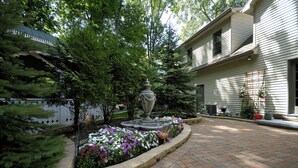 Une cour avec une fontaine, des parterres de fleurs et un chemin de briques menant à une maison dotée d’un porche.