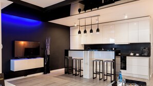 Een moderne keuken met witte kasten, zwarte aanrechtbladen en hanglampen. Er is een eethoek met barstoelen en een aan de muur bevestigde televisie.