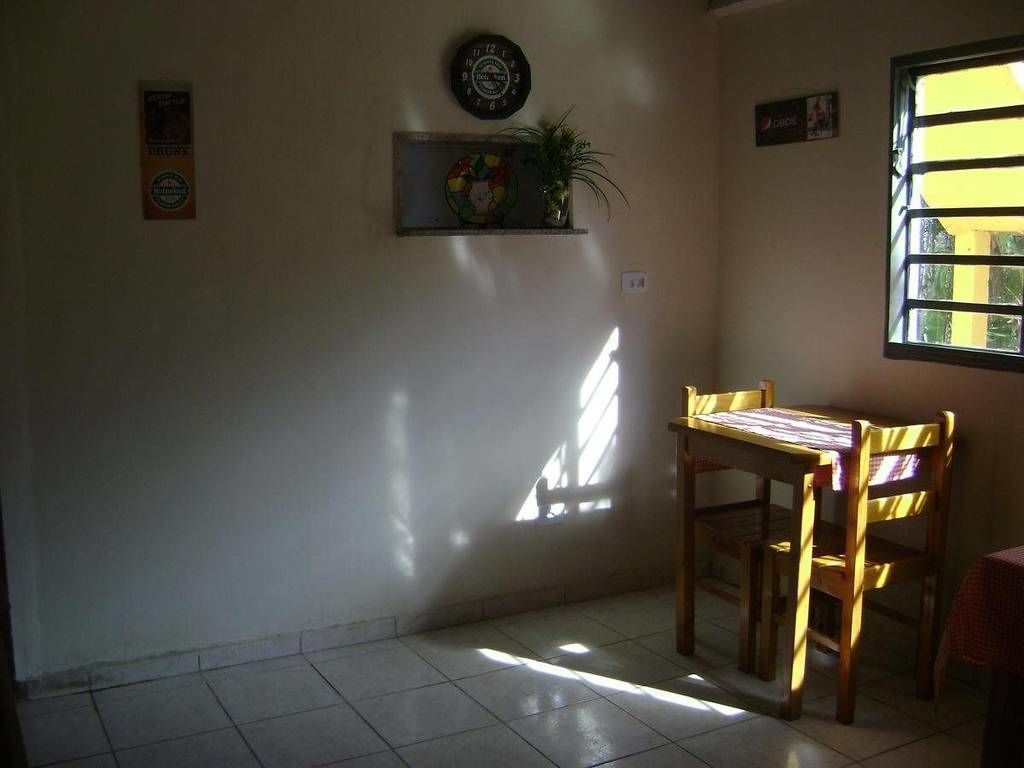 Una habitación con suelo de baldosas, una silla de madera, una mesita, un reloj de pared, una estantería con una planta y una ventana con persianas.