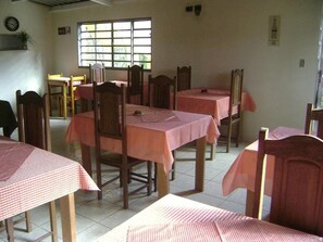 A dining area with multiple tables and chairs, a window with blinds, and a clock on the wall. - Pousada dos Tucanos (Sao Thome das Letras)