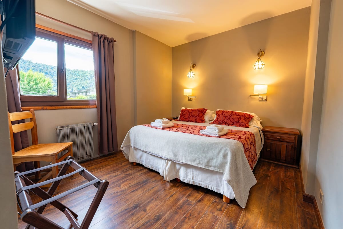 A bedroom with a wooden floor, a bed with red and white bedding, two wall-mounted lights, a window with curtains, and a chair.