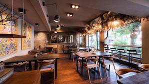Interior de restaurante con mesas y sillas de madera, luces colgantes y ventanas amplias.
