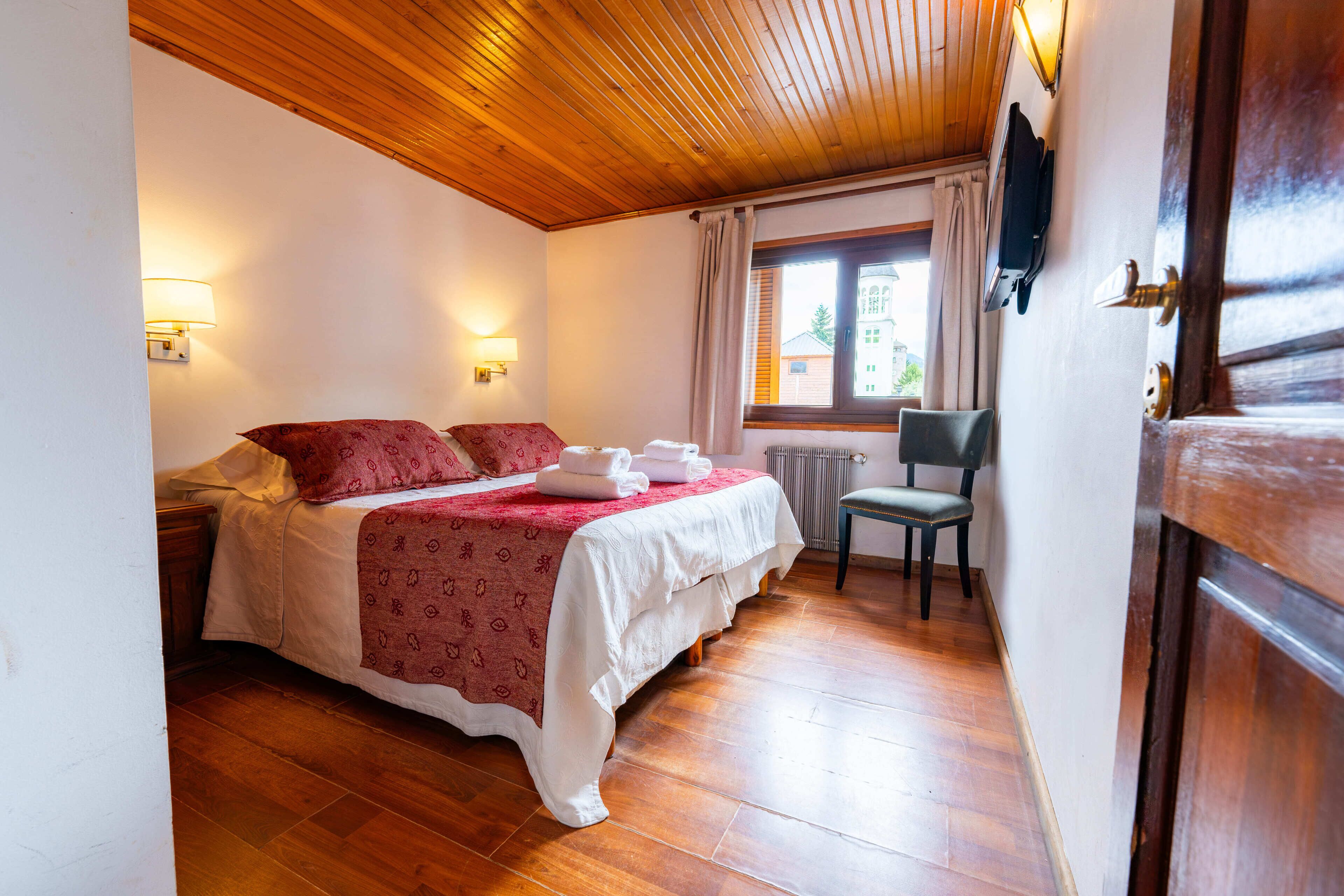 Una camera da letto con soffitto in legno, un letto con copripiumino rosso e bianco, due lampade ai comodini, una sedia e una finestra con tende.