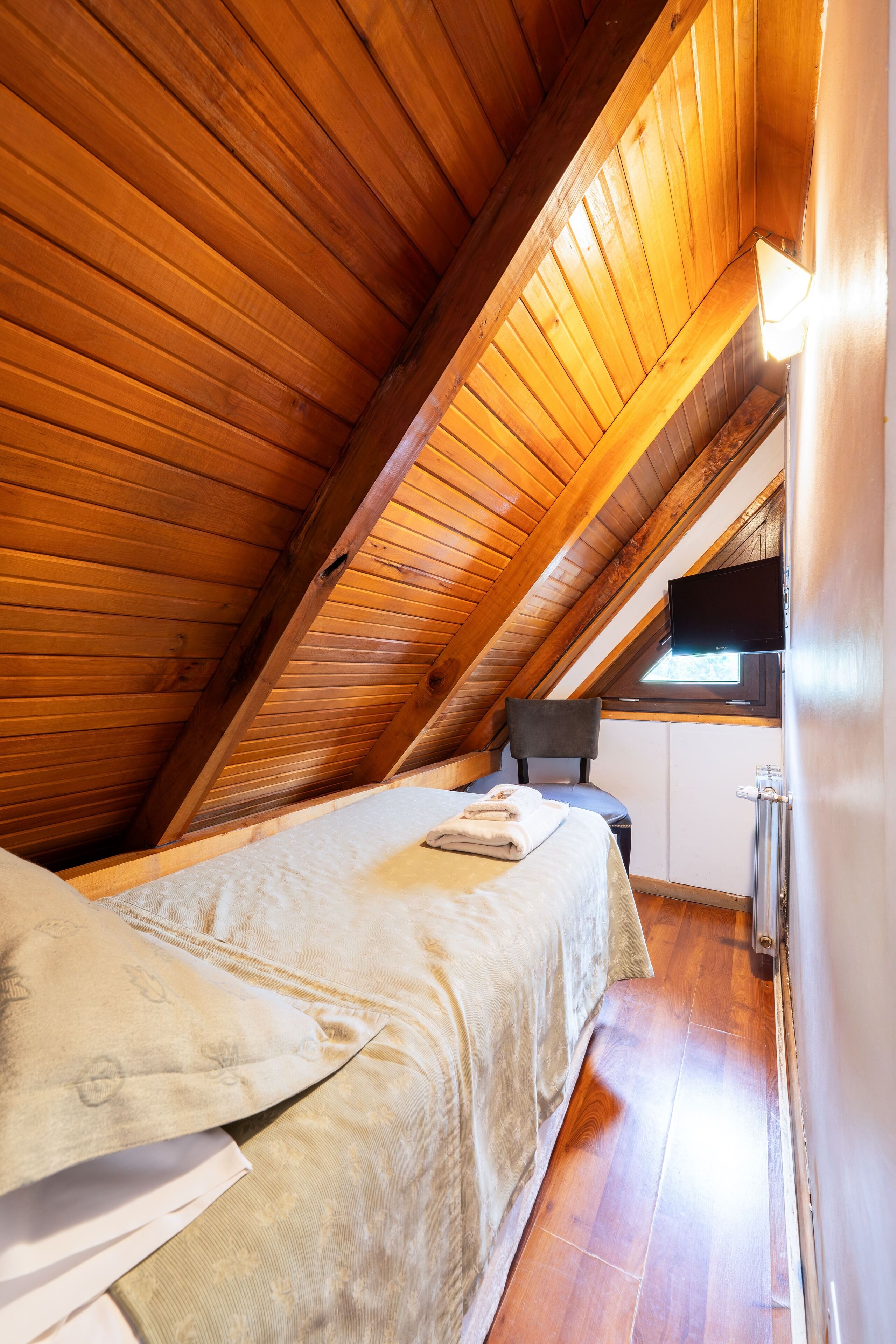 Un dormitorio con techo inclinado, cama, silla, televisor y ventana.