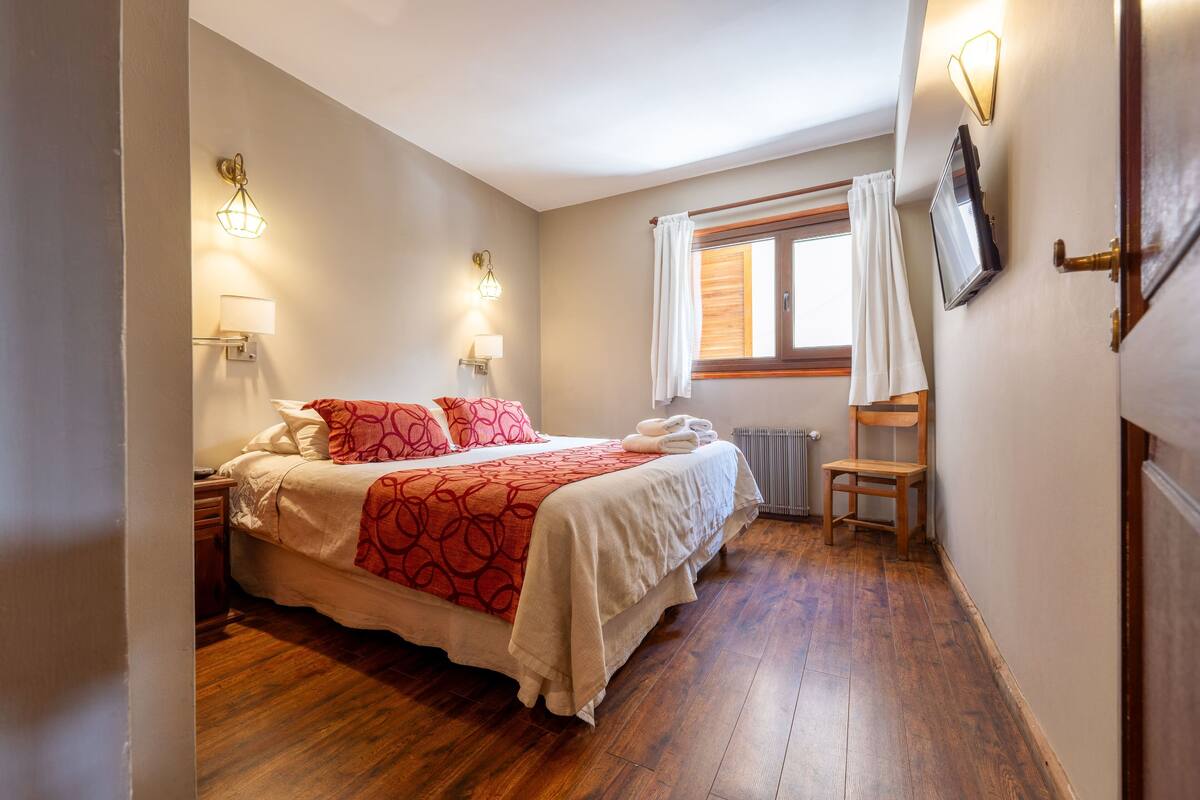 A bedroom with a bed, bedside lamps, a window with curtains, a radiator, and a wooden floor.
