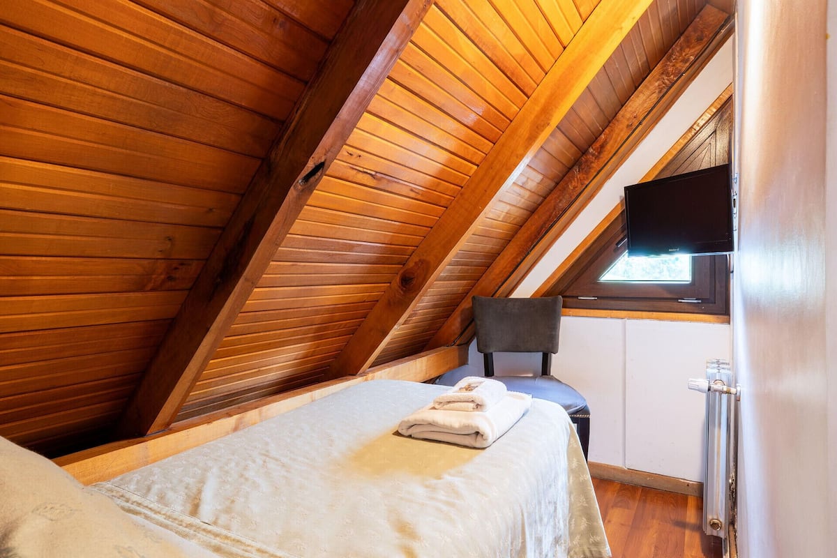 A bedroom with a sloped ceiling, a bed, a chair, a television, and a window.