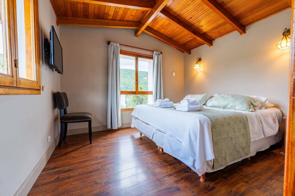 A bedroom with a wooden ceiling, a bed with white bedding, a chair, a window with curtains, and a wall-mounted TV.