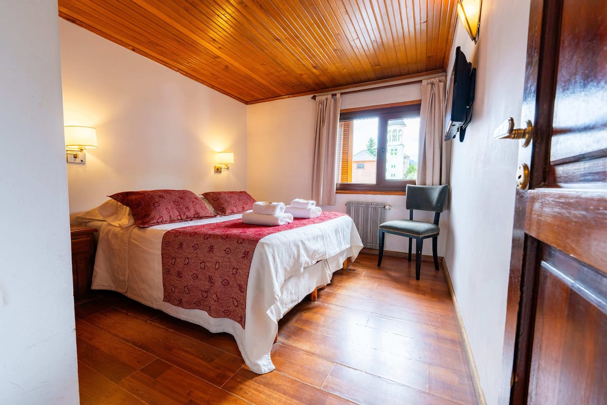 A bedroom with a wooden ceiling, a bed with a red and white bedspread, two bedside lamps, a chair, and a window with curtains.