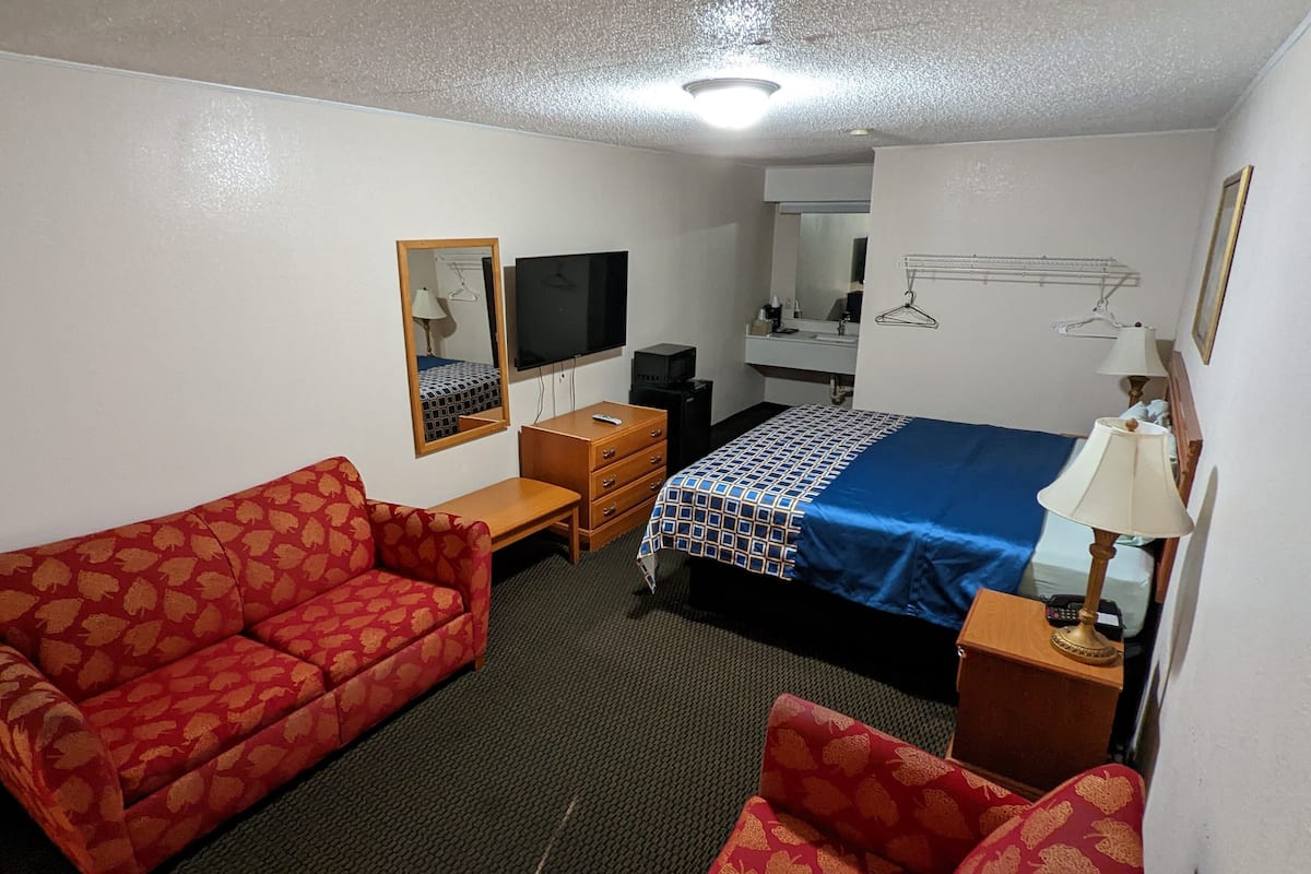A hotel room with a red patterned sofa, a bed with a blue and white patterned bedspread, a wooden nightstand with a lamp, a small wooden dresser, a TV, and a mirror on the wall.