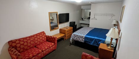 A hotel room with a red patterned sofa, a bed with a blue and white patterned bedspread, a wooden nightstand with a lamp, a small wooden dresser, a TV, and a mirror on the wall.