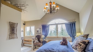 Une chambre à coucher avec un grand lit, une tête de lit en bois, un lustre, une fenêtre avec des rideaux et un ours en peluche sur le lit.