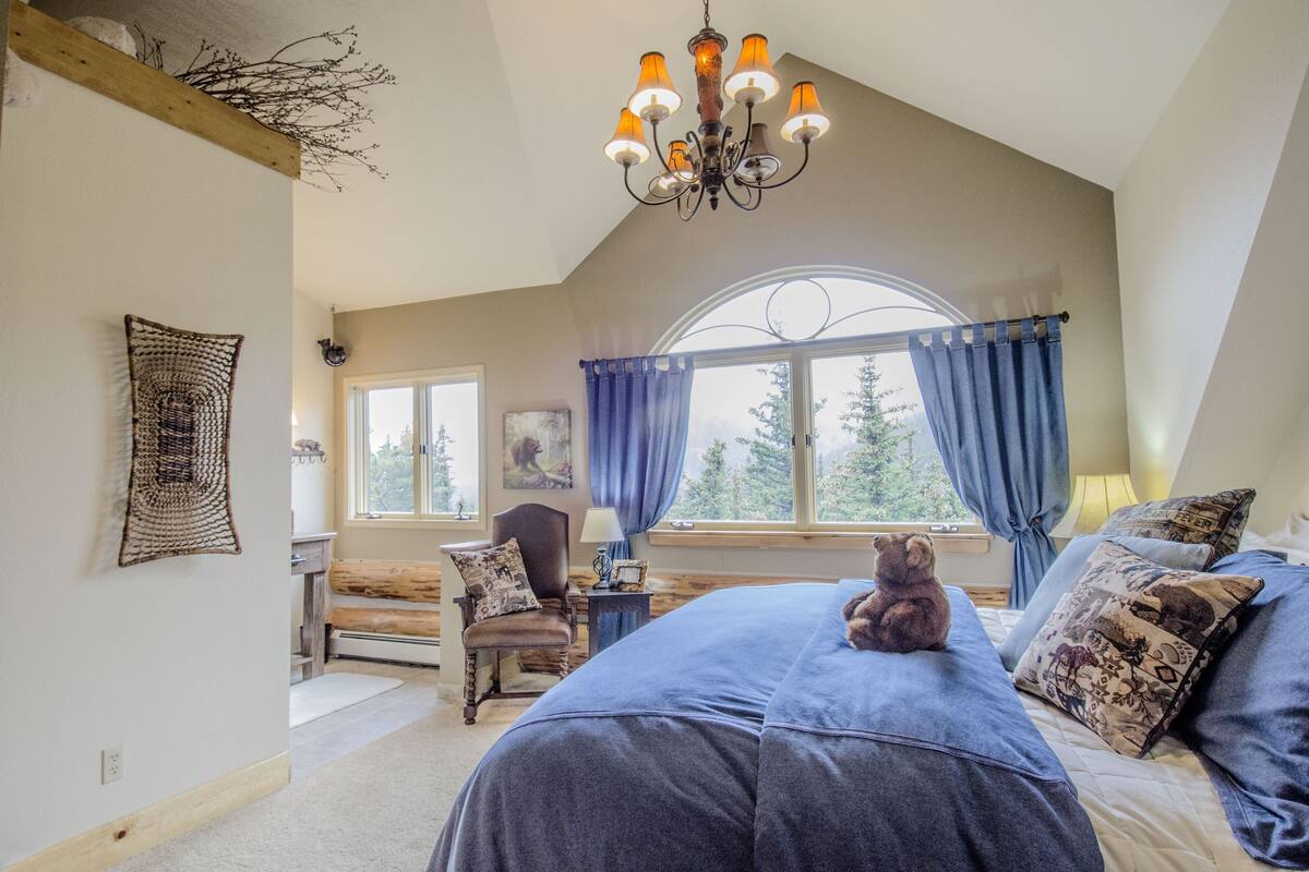 A bedroom with a large bed, a wooden headboard, a chandelier, a window with curtains, and a teddy bear on the bed.