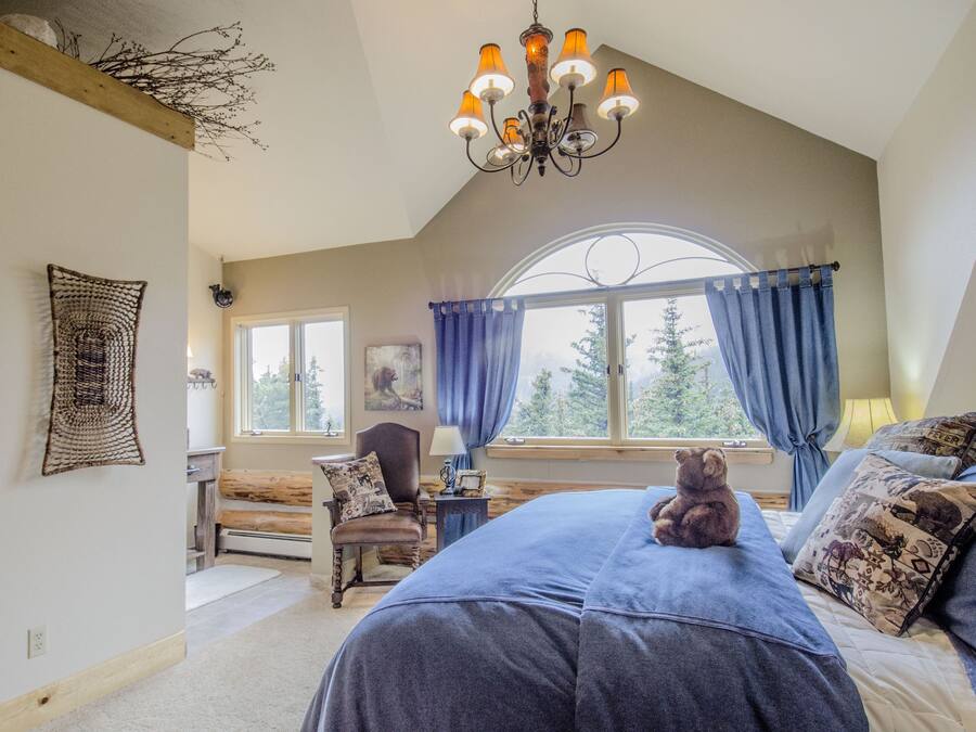 A bedroom with a large bed, a wooden headboard, a chandelier, a window with curtains, and a teddy bear on the bed.