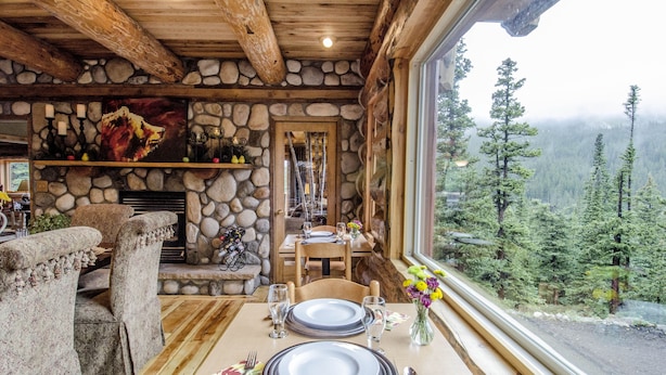 Un coin repas agrémenté d’une cheminée en pierre, de poutres apparentes en bois et d’une table dressée pour deux personnes, avec assiettes, verres et couverts.