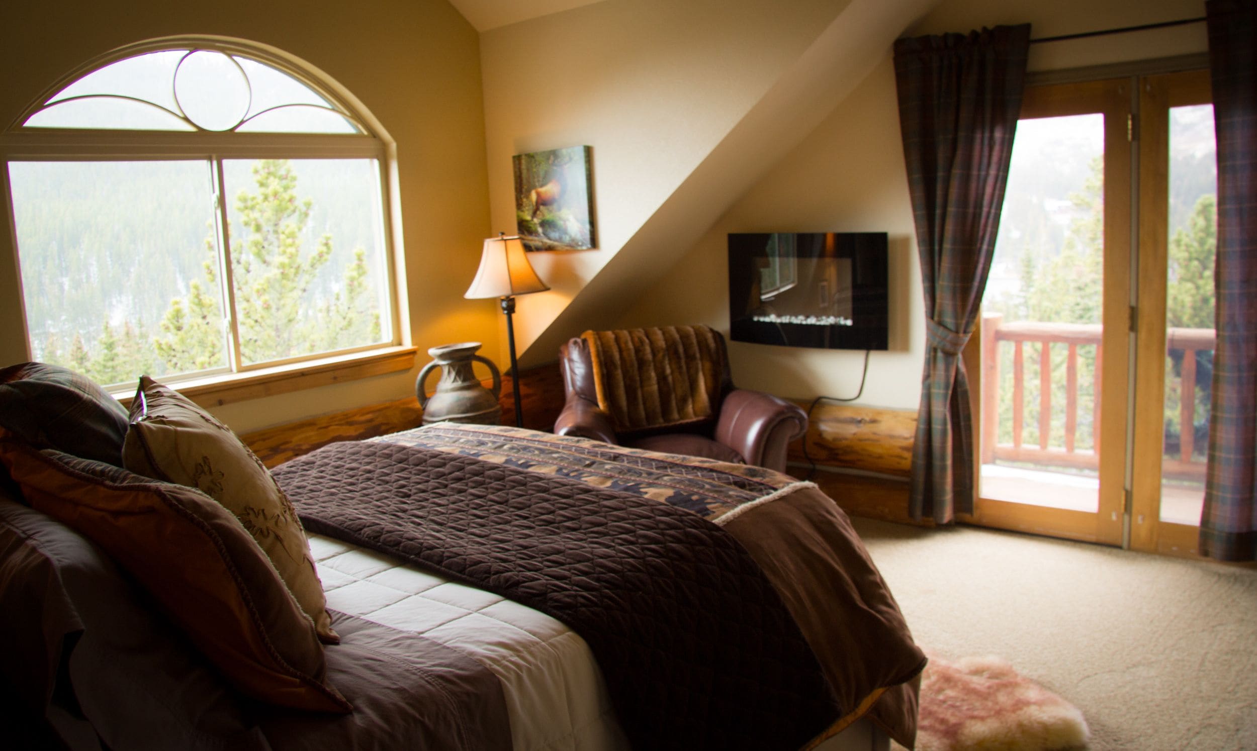 A bedroom with a large bed, a TV mounted on the wall, a window with a view of trees, and a chair by the window.