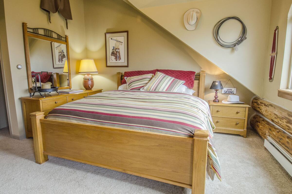 A bedroom with a wooden bed, a nightstand with a lamp, a dresser, a mirror, and a wall-mounted ski sign.