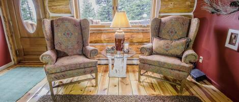 A cozy room with two patterned armchairs, a small table with a lamp, and a view of trees through large windows.