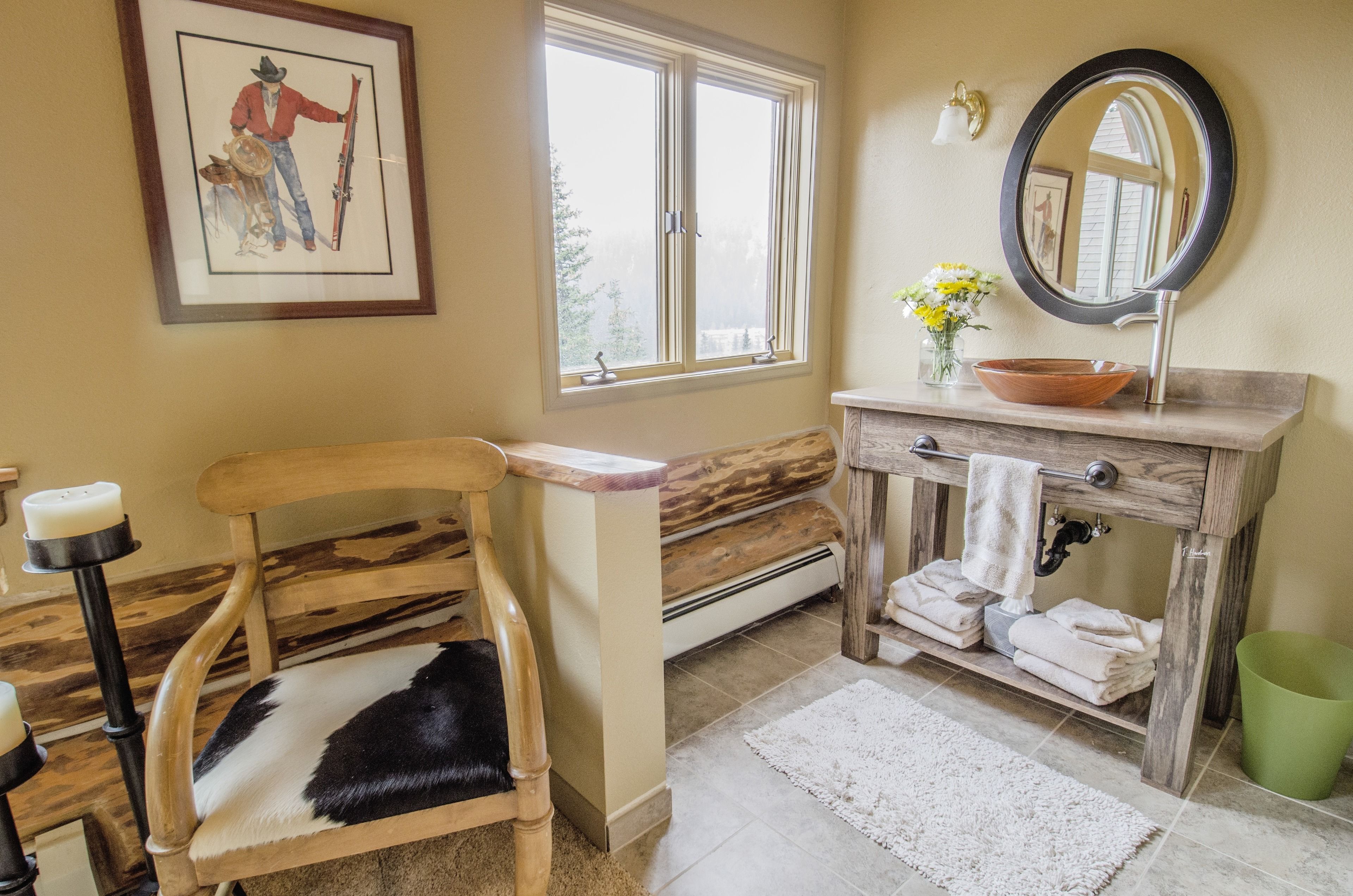 Une salle de bain avec une chaise en bois, une grande fenêtre, un lavabo avec un miroir et un vase rempli de fleurs.