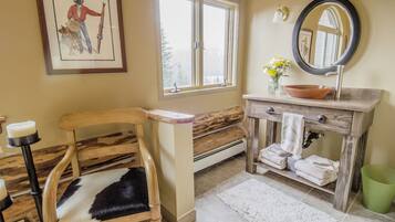 Une salle de bain avec une chaise en bois, une grande fenêtre, un lavabo avec un miroir et un vase rempli de fleurs.