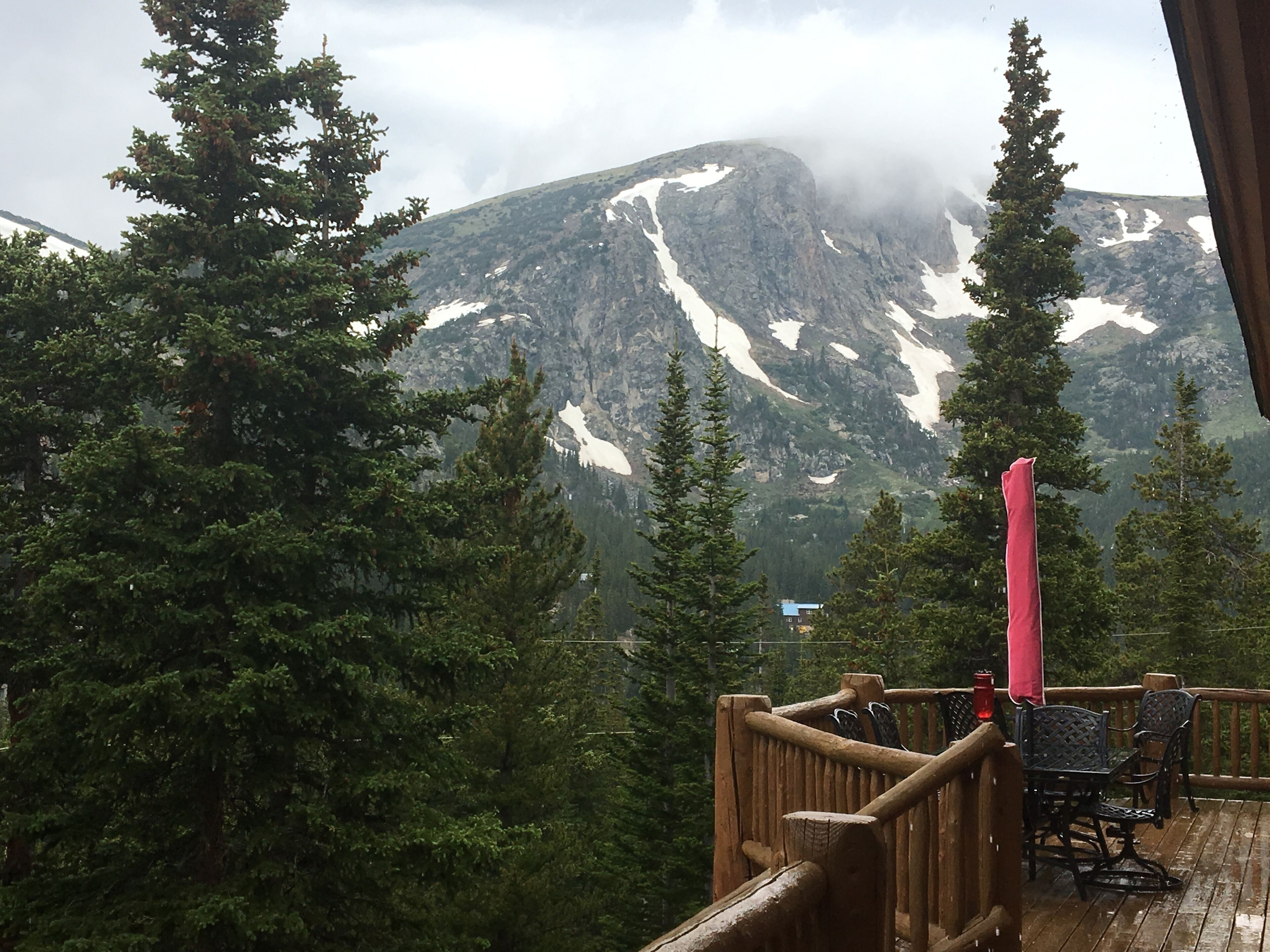 A wooden deck with outdoor seating, overlooking a mountainous landscape with snow patches and dense forest.